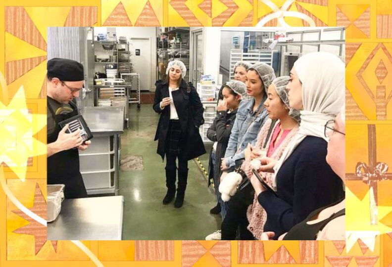 A photograph of the interior of a chocolatier's kitchen. An employee is doing a tour for a group of people who are all wearing head coverings and looking intently at their tour guide, who is describing the making of chocolate treats. The photo is set against a festive gold background accented with images of stars, gift boxes and ribbons.