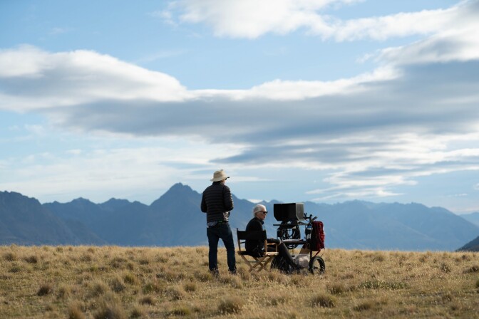 Two people, one sitting at a camera in a directors chair, the other standing in a cowboy hat, in the middle of a field with dry grass and mountain in the background.