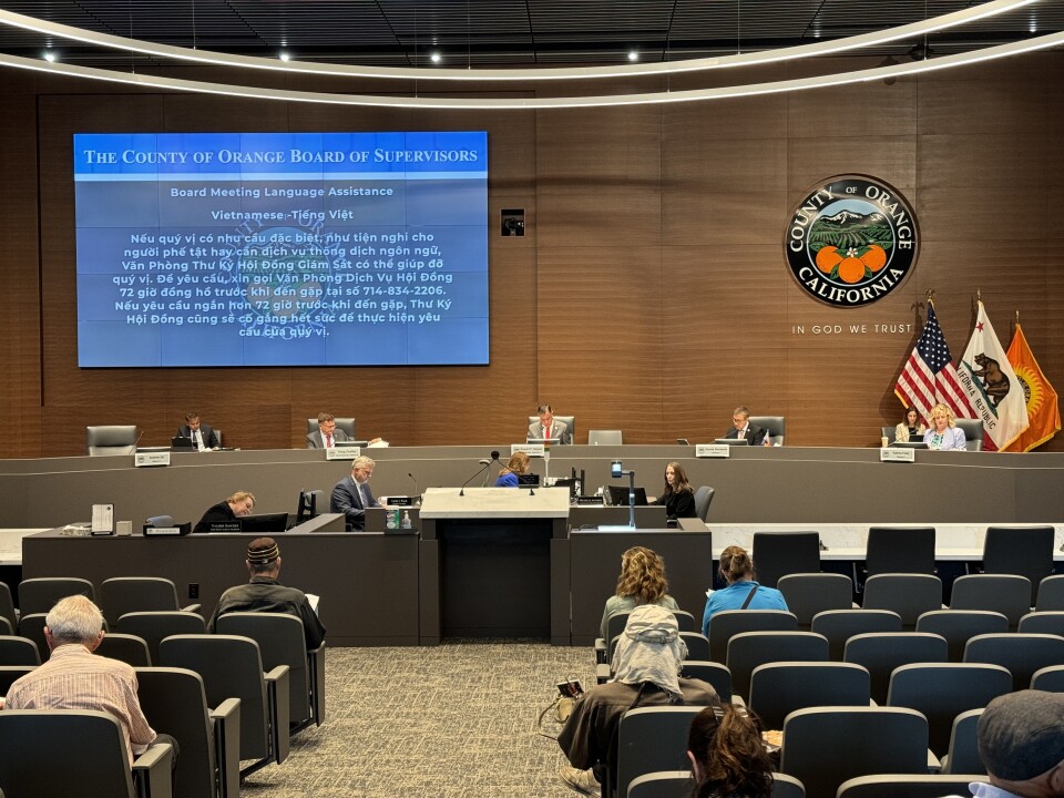 The seal for Orange County is on the wall of a board meeting. On a screen is information on how to access Vietnamese language assistance. 