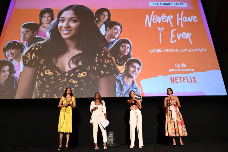 Four Asian American women in formal dress stand on a stage with a giant screen behind them that displays photos of the cast of the Netflix show "Never Have I Ever."