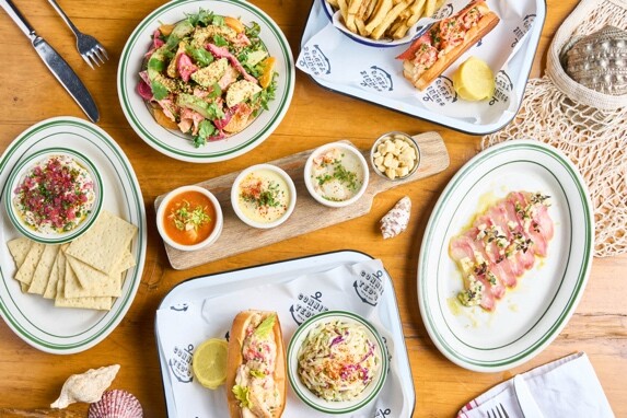 Bright overhead shot of a seafood meal including lobster rolls, a salad, fries, three small dips on a wooden board, crackers, a bowl of coleslaw, and a plate of light pink crudo arranged on nautical-themed plates.