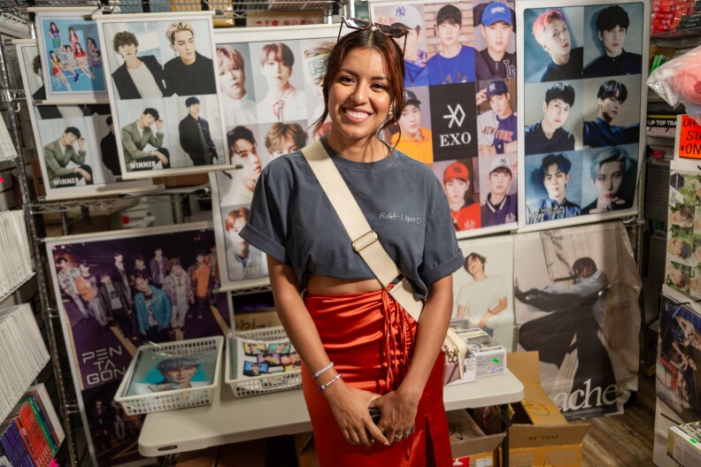 A female presenting person stands in front of a wall of K-pop posters. They are wearing a grey tshirt and orange skirt with a bag slung across their body.