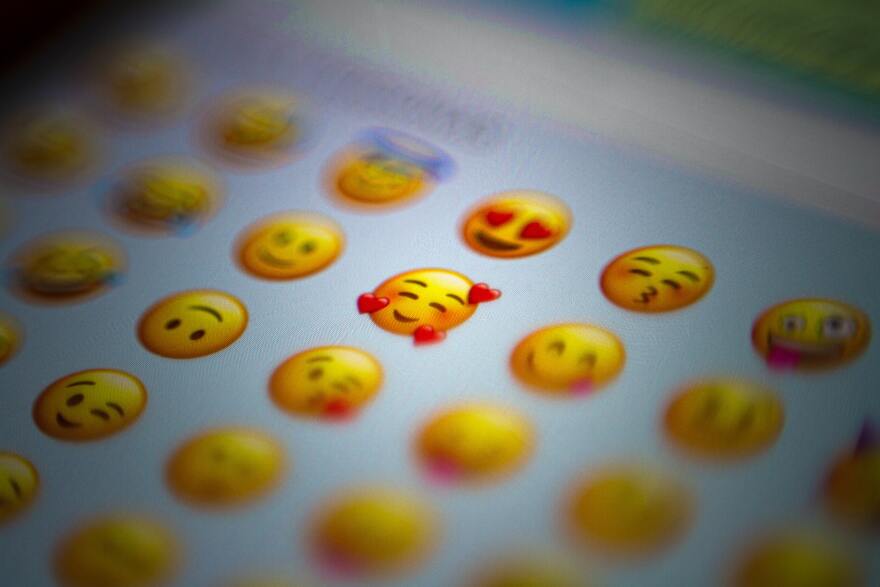 Closeup of a collection of upbeat yellow face emojis on a phone screen, with focus on a smiling, blushing face surrounded by hearts.