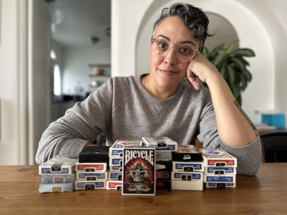 A person with medium-light skin tone, short hair and glasses sits at a wooden table with many decks of playing cards in boxes stacked on it.