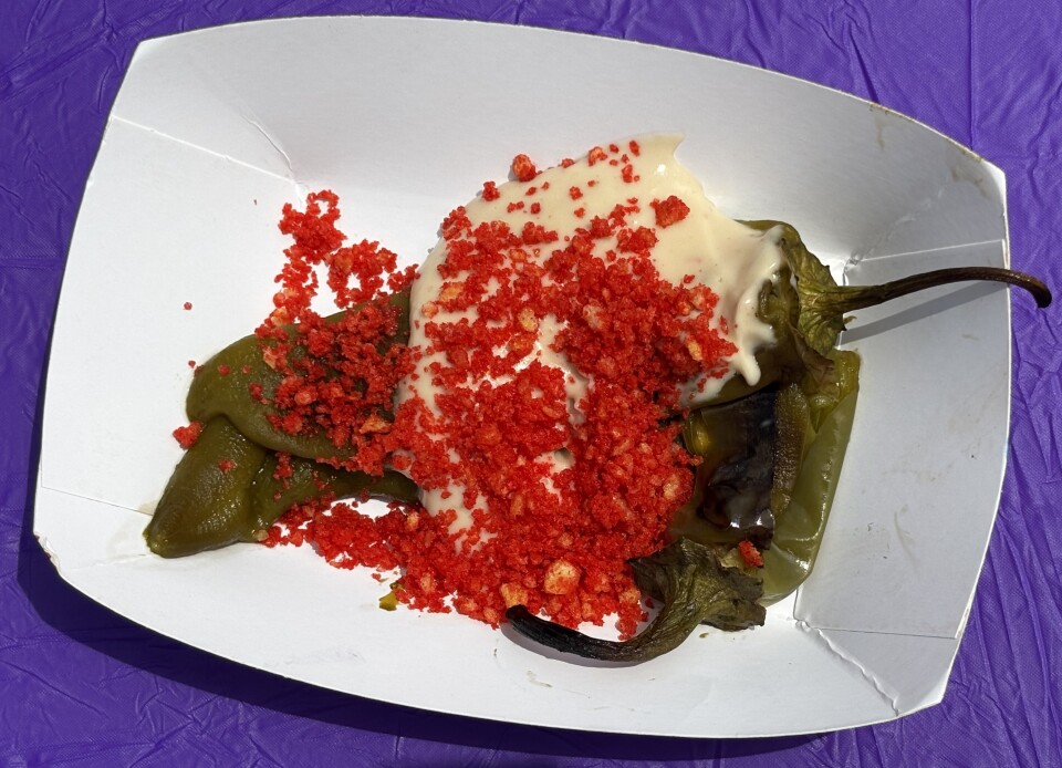 Roasted chile pepper smothered in a creamy, melted cheese sauce and generously topped with crushed Flamin’ Hot Cheetos, served in a classic paper food tray set against a purple tablecloth.
