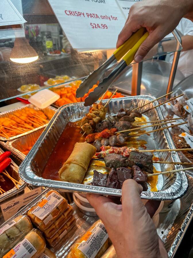 A pair of light brown hands holds metal tongs over an aluminum tray containing various skewered meats next to a counter that's filled with different types of food with a glass barrier and a white heat lamp.