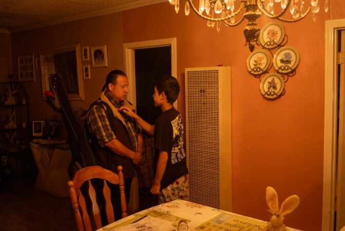 An older man and a teenage boy stand in a dimly lit living room.