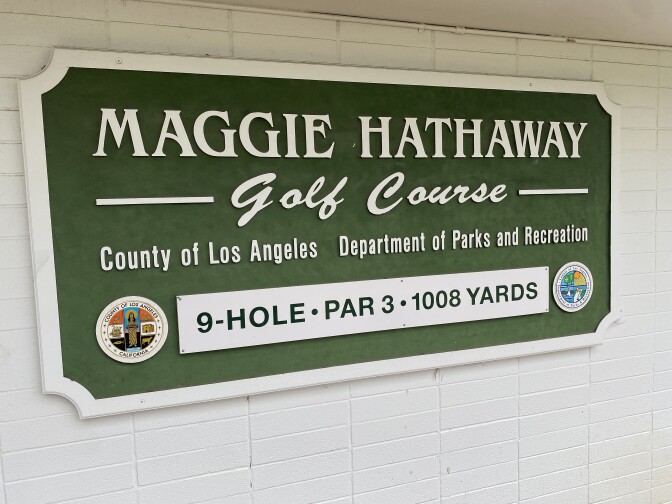 A sign in white lettering on a green background says "Maggie Hathaway Golf Course — County of Los Angeles — Department of Parks and Recreation." At the bottom of the sign in black lettering on a white background it says, "9-hole par 3 - 1,008 yards."