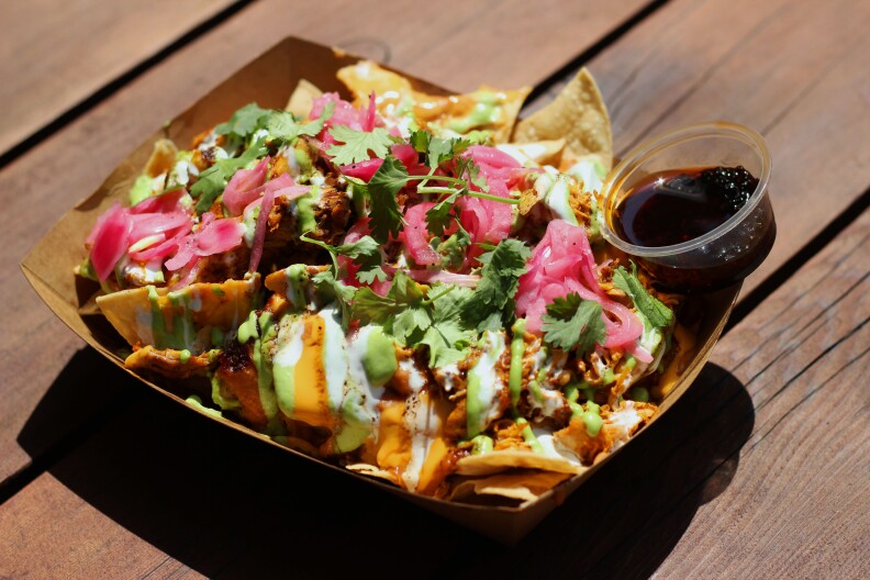 a brown cardboard food container holds a pile of tortilla chips, meat, pickled onions and white sauce