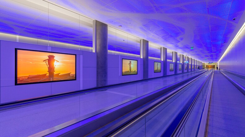 A rendering of a moving walkway illuminated in purple light. The walkway move from the bottom of the image to the distance. Images of the air traffic control in a golden sun set are on the wall between dark grey pillars.