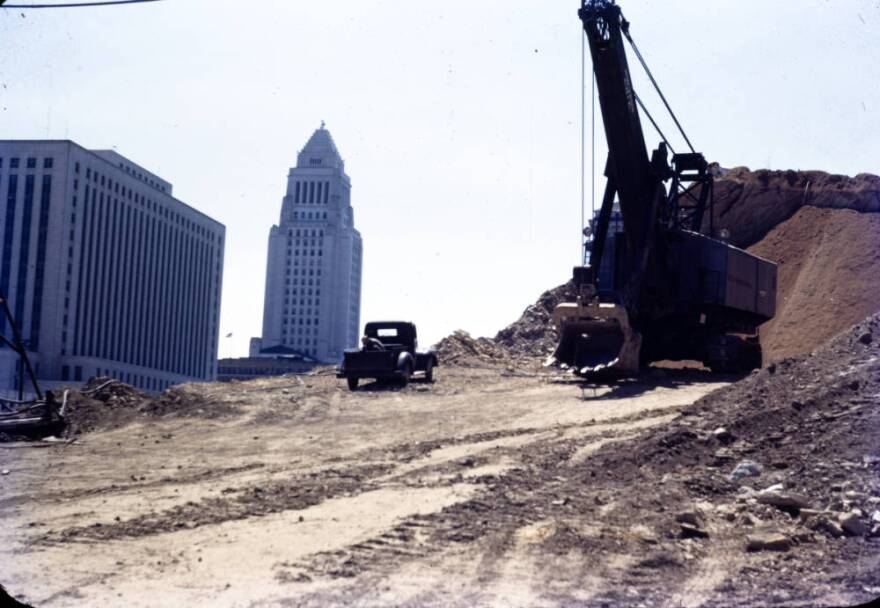 Looking southeast from Fort Moore Hill during its removal. The area is dirt with a car and an excavator machine. In the background is the U.S. Post Office, courthouse and City Hall.