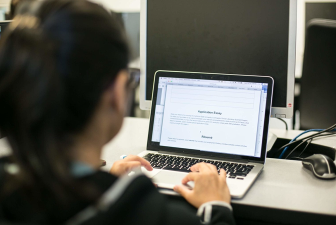 A person is sitting behind a laptop and typing with the screen reading "Application Essay".
