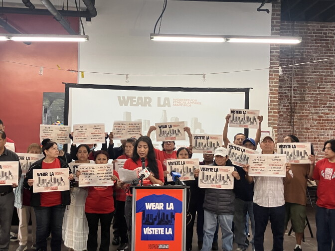 A group of people, some in red shirts, stand behind a podium holding signs that say "Wear LA" and "Visiete LA" with images of the Los Angeles skyline. One woman with a medium skin tone and long dark hair speaks at a microphone. There is a screen behind them that also says "Wear LA."