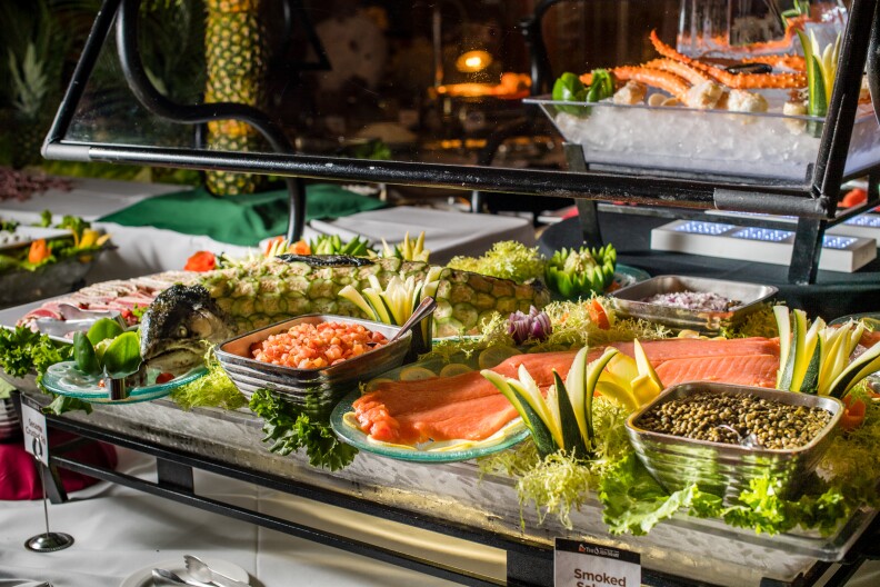 A buffet table laid out with a variety of seafood options, included lox, smoked fish salad (that is actually shaped like a sea serpent, and covered in thinly shaved cucumber "scales"). And off to the side is a platter filled with slices of seared tuna, as well as crab legs on crushed ice. The table also includes the traditional accents, such as diced onions and capers.