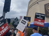 People holding signs that say "writers guild of america on strike" stand in front of a sign on the wall with the WB logo on it, Gate 4