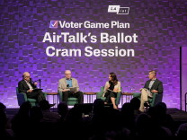 A photograph of a crowded, darkend room: The stage is dramatically lit in purple and there are four people sitting on stage in green club chairs, holding microphones.