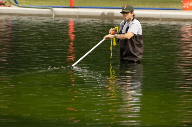 a man in a hat and waders stands waist deep in a body of green water and holds a long pole