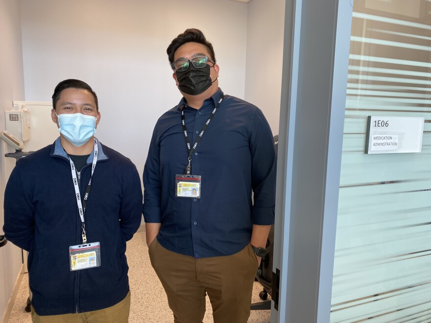 Nurse Quione Rodriguez and nurse practitioner Don Robeniol  stand side by side in a doorway to one of the medication administration rooms. They both wear face masks, blue button up shirts and tan slacks. 
