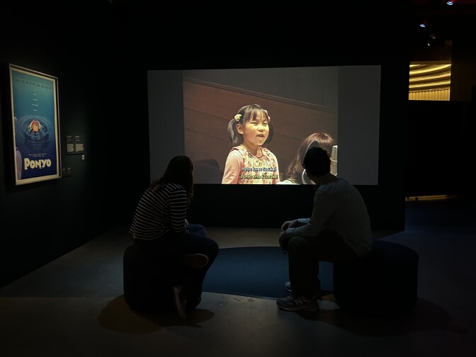 Two people are sitting on small chairs in the middle of a dark room, with their backs towards the camera. They're facing a screen playing a clip of a young Japanese girl. A blue movie poster for the film Ponyo is framed on the left wall.