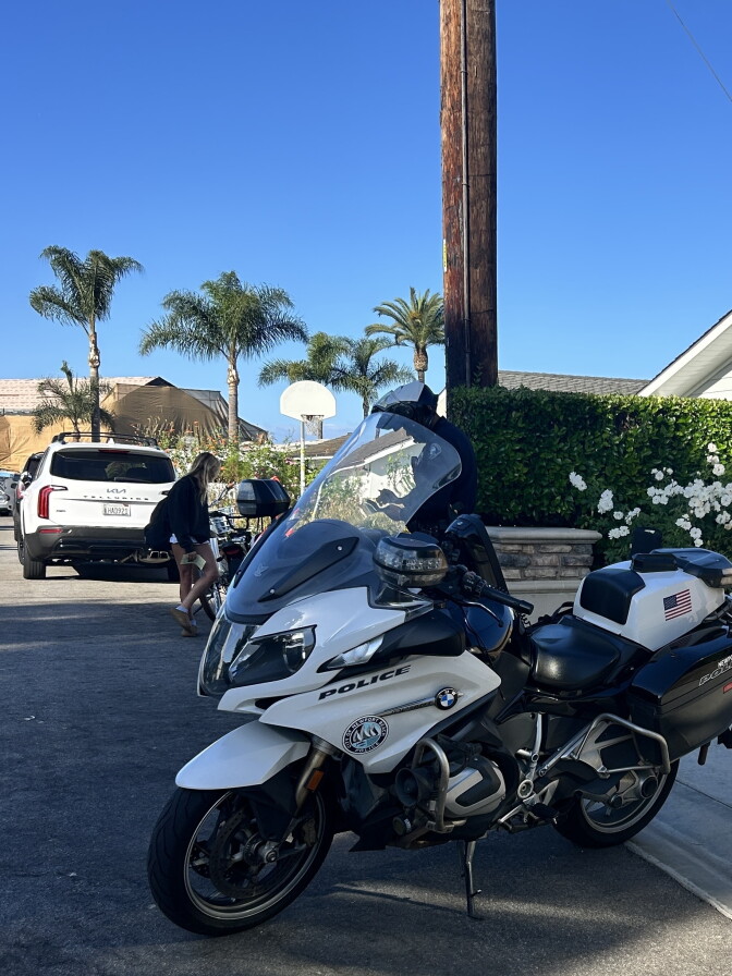 A Newport Beach police motorcycle is parked on a driveway lined with palm trees and a hedge with white flowers. A girl standing next to an e-bike is visible in the background.