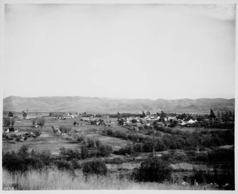 A black and white photo of open land with trees and a handful of buildings in the background.