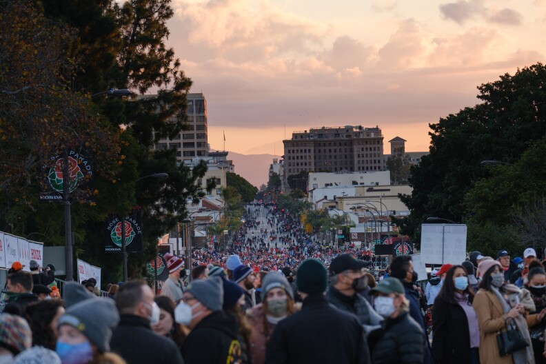 Hundreds of people walk on the streets in the early hours before the Rose Parade under a dawn sky.