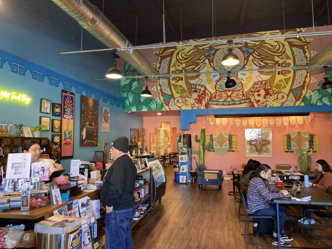 An interior view of a brightly colored bookstore with dark wood floors, blue and orange walls, and a large indigenous Mexican mural at the center. At the right, two women and three men sit at a table working on their laptops. At left a man with a black sweatshirt and black beanie speaks to a woman behind a counter with dark hair.
