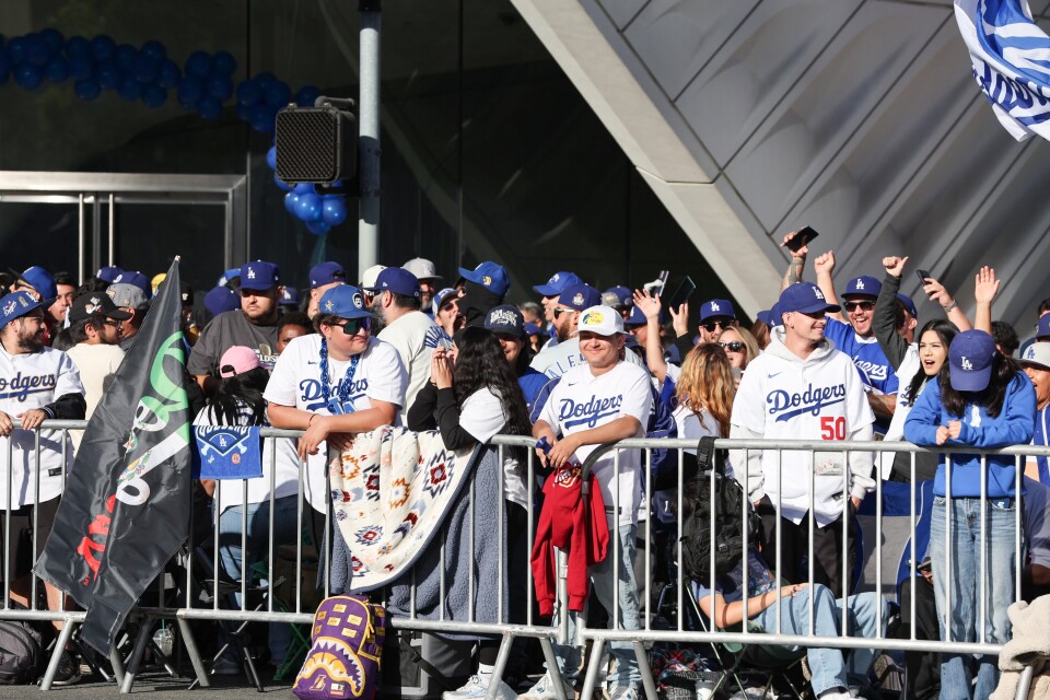 A group of fans wearing Dodgers gear wait behind metal barricades on a street. 