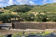 A wall with scaffolding stands in front of a hillside lined with trees and grass.  