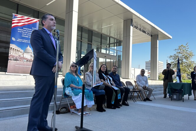 A middle-aged man with light brown skin and short graying hair wears a blue suit and speaks at a podium in front of a white rectangular government building. Five other people sit in chairs beside him, watching (three women, two men). In the background is blue sky. 
