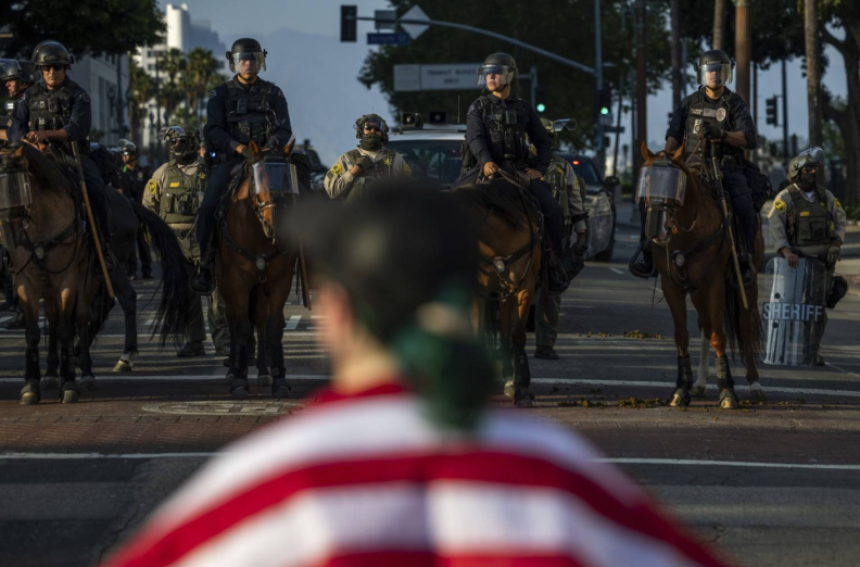 A person, out of focus in the foreground, stands on a street in front of a row of police officers on horses and some standing behind them.