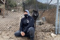 Wolf Connection team member Edward Amaya sits with hands clasped together. He wears a black jacket and grey hoodie. Beside him, behind a fence, sits his buddy Kenai, a black and brown male wolf who lives on the ranch. 