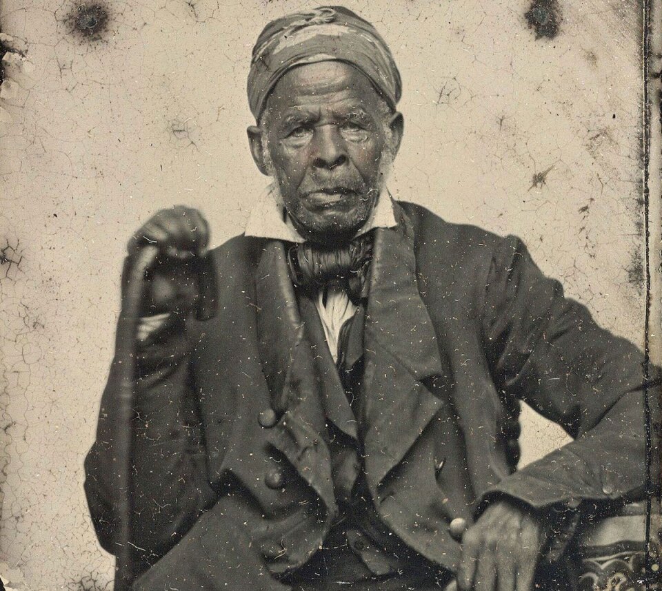 An antique portrait of Omar Ibn Said wearing a suit, resting on a cane and wearing a head scarf.