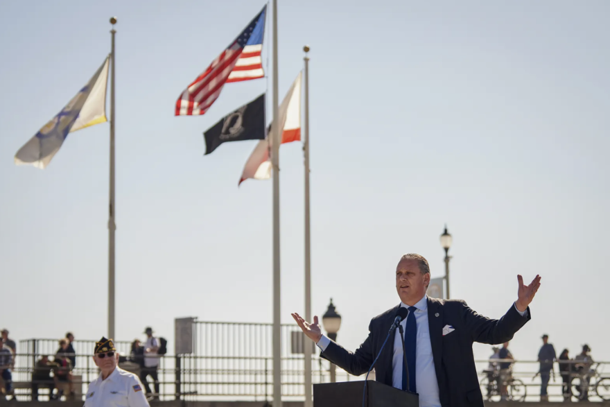 At the lower right of this images, a white man in a suit has his arms outstretched as he speaks into a microphone. Behind him are various flags. It's during the day.