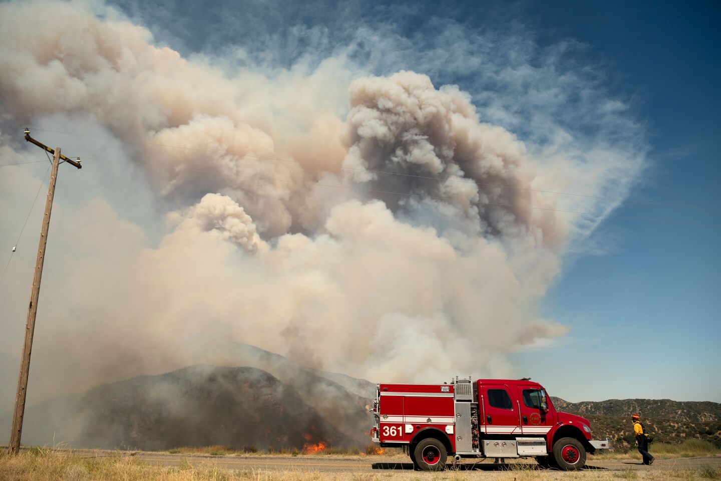 A massive gray and white cloud rises from the hills spreading ash near a bright red firetruck. One fighter, in a yellow shirt and dark overalls, stands to the right. A single powerline pole is at the left.