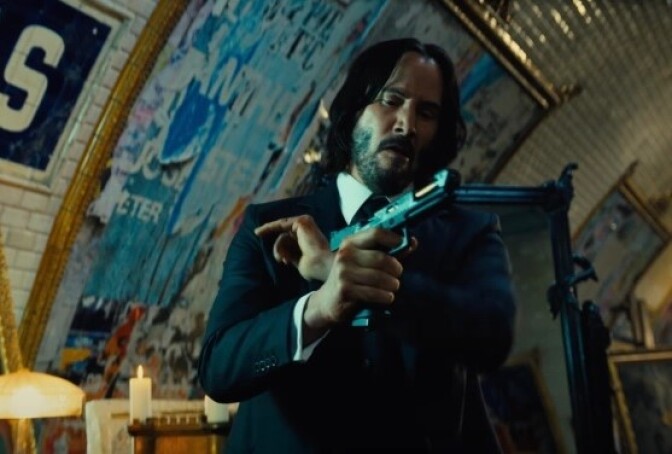 Keanu Reeves stands in a room with white tiled walls similar to a subway. There are pictures hanging on the walls that curve into a domed ceiling. Reeves wears a black suit with a  white undershirt. He holds a gun in his right hand. His other hand rest against his chest. He seems to be cocking the teh gun getting it ready to shoot. 