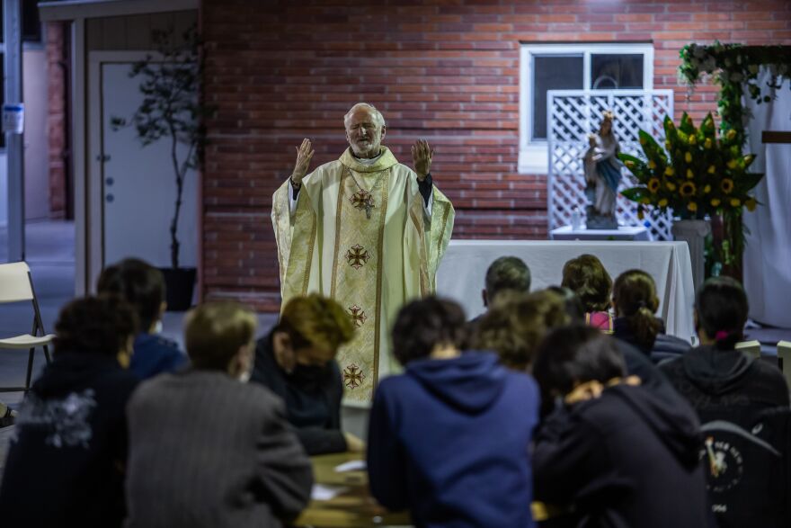 A man with gray hair and a beard wears a golden robe with crosses on it as he raises his hands in front of people with bowed heads.