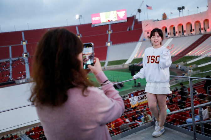 at an outdoor sports event, two femme presenting people stand taking a photo. Person in the foreground holds an iphone camera, person in the background is wearing a USC sweatshirt and poses