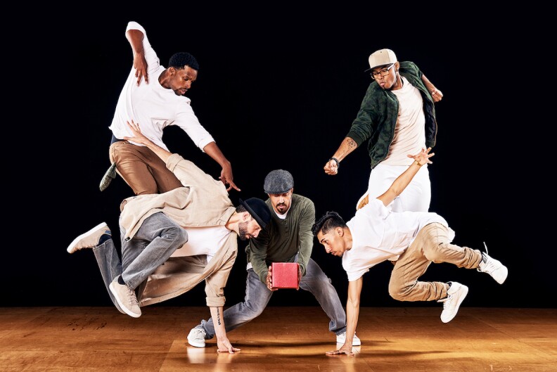 four men jump in the air in various dance poses while a fifth man, in the middle, holds a small box