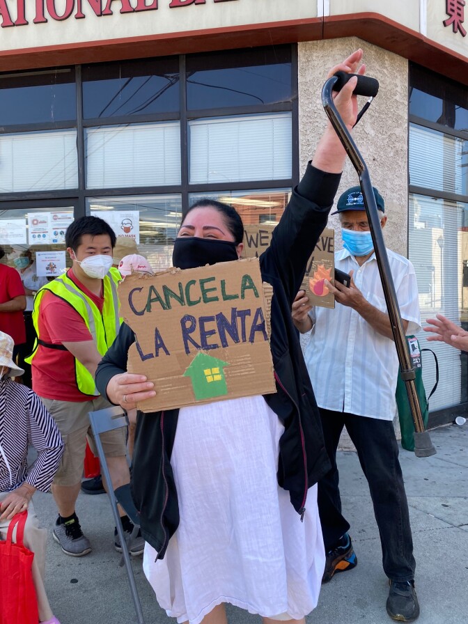 Ady Carrillo stands in front of Eastern International Bank with one arm raised and the other holding a sign that says "cancela la renta."