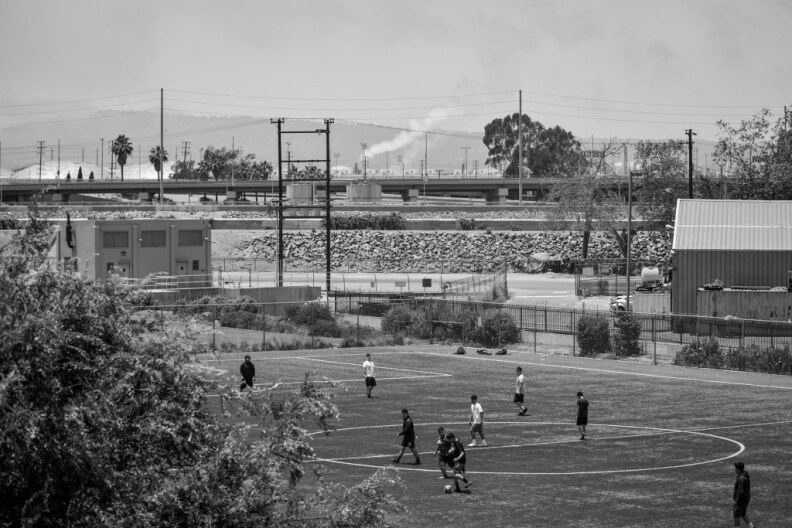 A wide shot view that's in black and white of the soccer field while people play on it. A tree is in the foreground.
