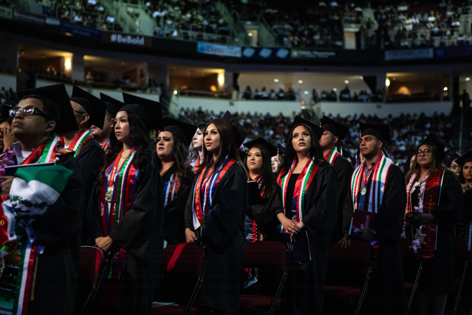 Rows of students inside an auditorium wear graduation attire.