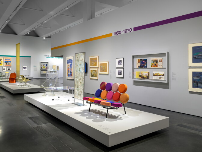 Gallery view of a 1950–1970 section of a design exhibition, with a sofa made of colorful round cushions on a white platform in the foreground, clear acrylic furniture behind it, and framed drawings, posters, and magazine pages on the walls.