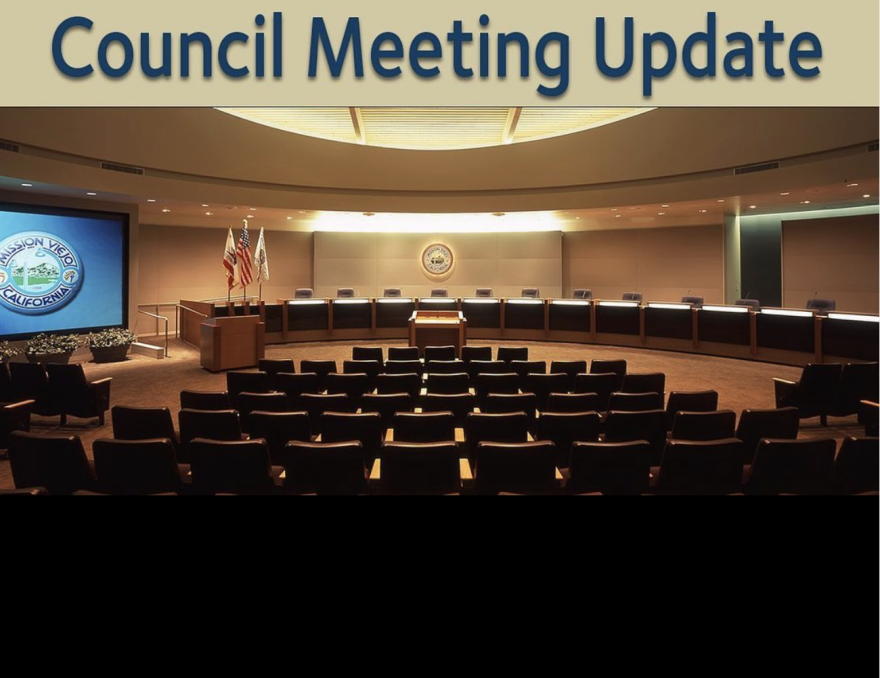 Photo is of an empty city council chambers with text at the top reading "City Council Update." On the left-hand side, there's a screen showing the image of a seal reading "Mission Viejo, California." Further toward the dais, there's a stand with the U.S. and California flag and another flag.