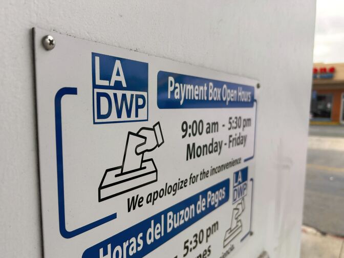 A close up of a payment drop box sign on the white wall of the LADWP building. It says in blue, black and white the LADWP payment box open hours with an icon of a hand putting an evelope into a box cutout. The box is open 9am to 5:30pm Monday through Friday. Below it has the words in Spanish.