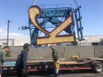 Three workers surround a flatbed truck, as a gigantic sign of the letter "K" -- tilted on its side -- is being lowered onto it