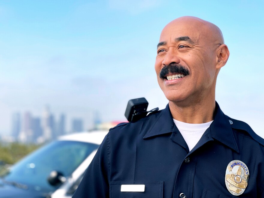 Cecil Rhambo in his LAX Police Chief uniform, looking off in the distance, with a police car behind him and the LA skyline in the far distance.