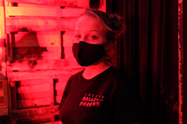 A woman stands in a wooden hallway flooded with red light. She wears a facemask and a black T-shirt with a red title across the chest that says LACHSA HALLS OF HORROR.