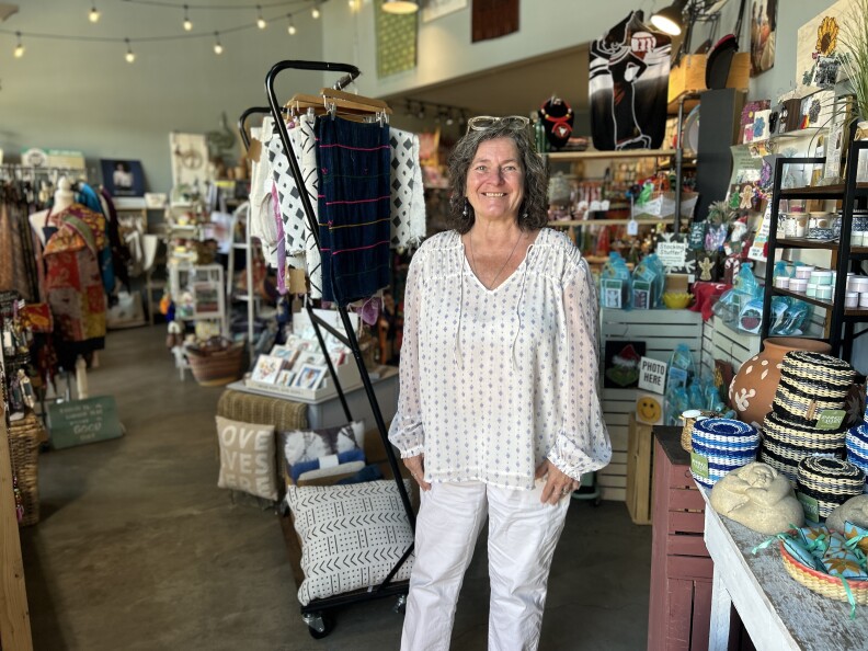 A white woman with short, gray curly hair smiles for the camera wearing a flowy white shirt and pants and clear glasses on her head. She is in a store surrounded by colorful goods.