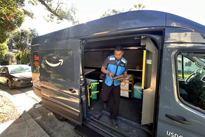 A man wearing black jeans a blue work vest over a t-shirt is stepping out of the back of a navy Amazon delivery van in a residential neighborhood. 